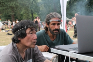 Zwei Personen mit „Stromabnehmer“ auf dem Drogeninfostand auf dem New Healing Festival 2017. Foto: Tomek (CC BY-SA 3.0 DE). Links im Bild ist der Entwickler dieser Technologie, Masahiro Kahata, zu sehen. Umgangssprachlich wird diese Technologie auch „Gehirnwellenstriptease“ genannt